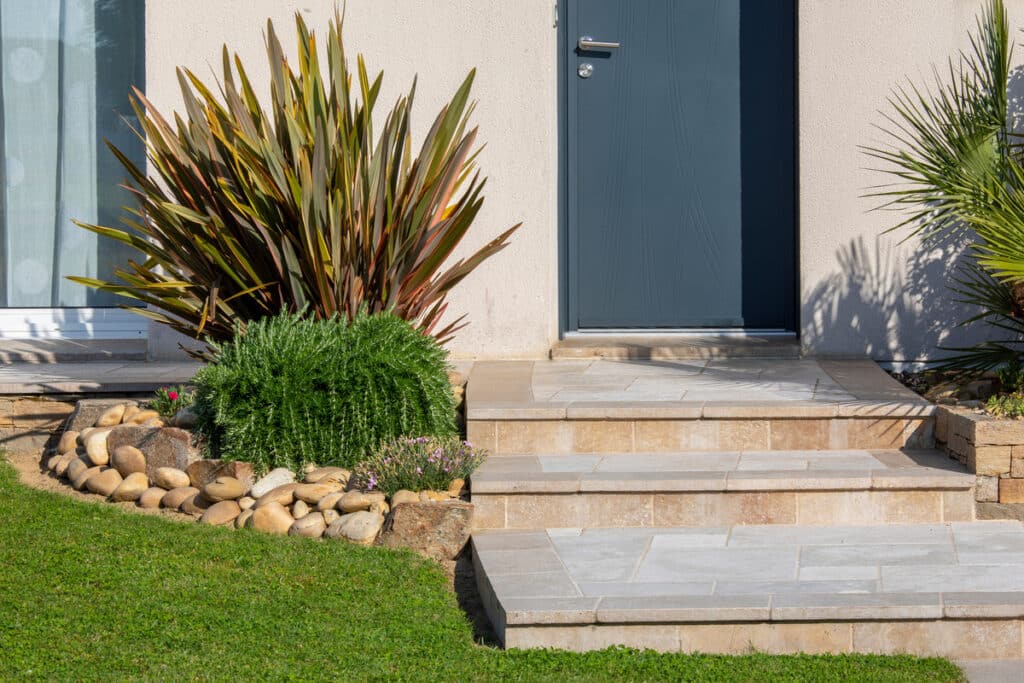 Escalier en pierre menant à une porte grise d'entrée de maison, flanqué de plantes et de rochers dans le jardin.