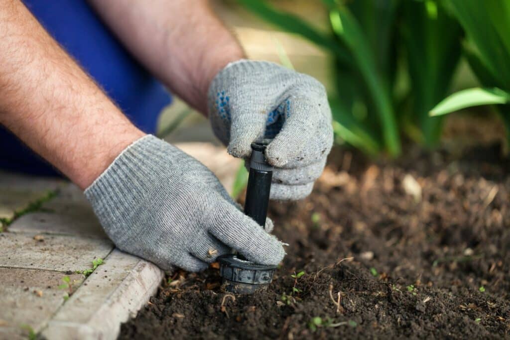 Mains gantées ajustant une tête d'arroseur dans le sol d'un jardin à côté d'une bordure.