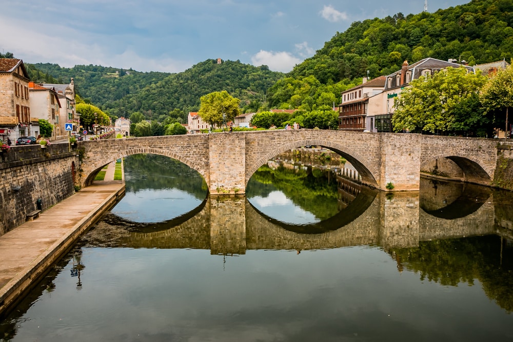 Pont en pierre à trois arches sur rivière, reflets dans l'eau, bordé par des bâtiments de ville et collines verdoyantes.